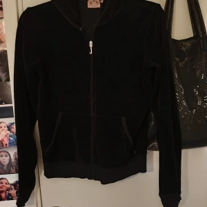 Svart velour hoodie Juicy Couture XL - Klassisk svart hoodie från Juicy Couture i mjuk velour med dragkedja och huva. Har två fickor fram och ikonisk J-dragkedja. Perfekt för en chill och trendig look, passar dig som gillar comfy vibes och streetstyle.