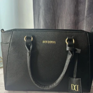 Svart handväska från Don Donna - Stilren svart handväska från Don Donna med guldfärgade detaljer och logotyp framtill. Väskan har både handtag och en avtagbar axelrem, samt praktiska fack och dragkedja inuti. Perfekt storlek för att få plats med allt du behöver och passar till många olika outfits.