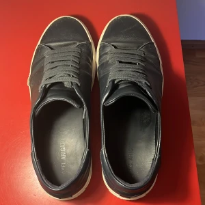 Mork blå sneakers från Axel Arigato - Stilrena svarta sneakers från Axel Arigato i skinn med vit sula och diskret logga i guld på sidan. Klassisk design med rund tå, snörning och lågt skaft. Perfekta för dig som gillar minimalistisk och clean stil.