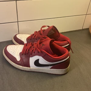 Nike Air Jordan 1 Low röd/vit - Säljer ett par Nike Air Jordan 1 Low i rött och vitt med svarta detaljer. Klassisk siluett med svart swoosh, perforerad tåbox och röda skosnören. Skorna har en låg profil och är tillverkade i skinn och syntet. Perfekta för dig som gillar streetwear och sneakers med attityd.