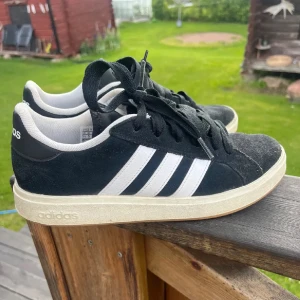Svarta Adidas sneakers med vita ränder - Snygga svarta Adidas sneakers med klassiska vita ränder på sidorna och vit sula. Skorna har snörning och är gjorda i mocka och syntetmaterial. Perfekta för en avslappnad och sportig stil.