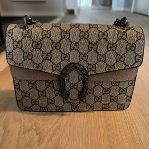 Fin axelväska  - Säljer en snygg axelväska  i beige och brunt. Väskan har en kedjerem i metall och ett dekorativt spänne framtill. Perfekt storlek för det viktigaste och ger en lyxig touch till din outfit.