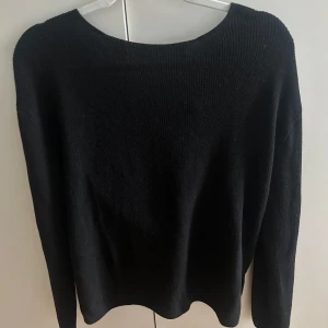 Svart tröja - Säljer en svart stickad tröja med snygg omlottdetalj i ryggen. Tröjan har lång ärm och en lite lösare passform, perfekt för dig som gillar en stilren men ändå trendig look. Passar till många olika outfits och är superenkel att matcha
