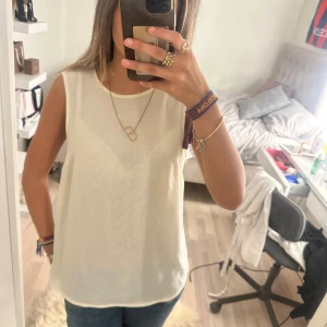 Vit plisserad blus i chiffong - Säljer en vit blus i tunn chiffong med plisserad rygg och knapp i nacken. Blusen är ärmlös och har en flowy, lätt transparent look som ger en chill vibe. Perfekt till jeans eller kjol för en fräsch och stilren outfit.