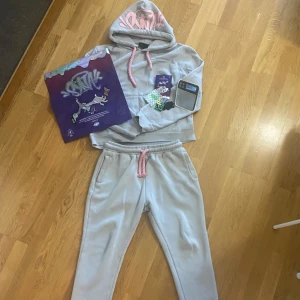 Grå Syna World Tracksuit - 🔥 Grå Syna World tracksuit 🔥 Supersnygg set med rosa snören och unikt rosa graffiti-tryck på huvan. Insidan är i mjuk fleece och tröjan har en stor, praktisk magficka framtill – perfekt för chill dagar och en avslappnad streetwear-look.  ✨ Detaljer som sticker ut: 	•	Broderad text på benet 	•	Matchande tryck på byxorna 	•	Storlek L men sitter perfekt även på M  👉 Ett måste för dig som vill ha både komfort och stil! 
