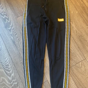 Svarta mjukisbyxor från Hugo Boss - Svarta mjukisbyxor från Hugo Boss med gula och vita loggor längs sidorna och en gul HUGO-logga på låret. Byxorna har dragsko i midjan och är i en slim passform. Perfekta för en chill och sportig look.