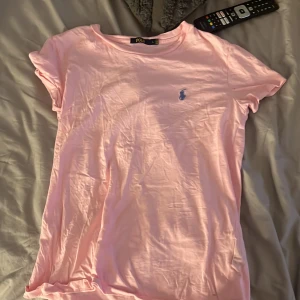 Ljusrosa t-shirt från Polo Ralph Lauren - Snygg ljusrosa t-shirt från Polo Ralph Lauren med klassisk blå broderad logga på bröstet. T-shirten har rund halsringning och korta ärmar. Perfekt för en chill och stilren look.