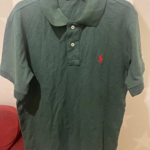 Grön pikétröja från Ralph Lauren - Snygg grön pikétröja från Ralph Lauren med klassisk krage och två vita knappar framtill. Röd broderad logga på bröstet ger en stilren touch. 