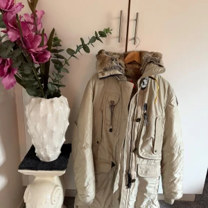 Beige Parajumpers parkas - Snygg beige parkas från Parajumpers med ikonisk gul rem vid kragen och patch på ärmen. Jackan har flera stora fickor, dragkedja och knappar framtill samt en huva med fuskpäls. Perfekt för kalla dagar och riktigt street.