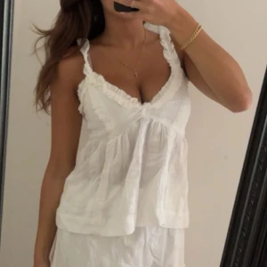 Vit volanglinnetopp med spetsdetaljer - Supersöt vit linnetopp med volangkant och spetsdetaljer längs v-ringningen. Luftig och lätt med smala axelband, perfekt för varma dagar. Ger en romantisk och somrig vibe.
