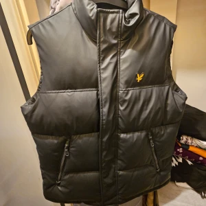 Svart dunväst från Lyle & Scott - Svart dunväst från Lyle & Scott med gul logga på bröstet. Västen har hög krage, två dragkedjefickor framtill och quiltad design. Perfekt för lager-på-lager och enkel att matcha med hoodies eller tröjor.