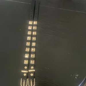 Valentino armband - Snyggt svart armband med fyrkantiga guldnitar i två rader och dubbla guldfärgade spännen. Perfekt accessoar för att ge outfiten en edgy vibe. Armbandet är i skinnimitation och har justerbara hål för bästa passform. Båda för 100 kr
