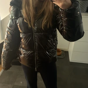 Svart glansig pufferjacka med huva - Jättensnygg trendig glans jacka från Nelly och använd några gånger men kommer ej tills användning (ser ut som ny). Perfekt nu till vintern! Denna jacka är oftast slutsåld på Nelly.