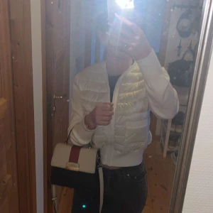 Catwalk handväska i svart och beige - Snygg handväska från Catwalk i beige och svart med en bred röd rand på locket. Väskan har guldfärgade detaljer, justerbar axelrem och ett stilrent, boxigt format. Tillverkad i syntetmaterial med struktur och logga framtill.