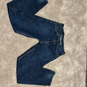 Carhartt Jeans 27/32 marlow pant - Dessa Carhartt-jeans kombinerar klassisk denimstil med slitstark kvalitet för vardag och arbete. Den djupa indigofärgen och de förstärkta sömmarna ger både tidlös look och lång hållbarhet. Med knappgylf, praktiska fickor och 100 % bomull erbjuder de komfort och funktionalitet. Perfekt val för dig som vill ha både stil och styrka i ett par jeans  inga stora skador men kan se lite slit på dom om man lägger ner tid.Dessa Carhartt-jeans kombinerar klassisk denimstil med slitstark kvalitet för vardag