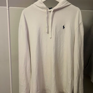 Vit hoodie från Polo Ralph Lauren - Vit hoodie från Polo Ralph Lauren i mjuk bomull med klassisk huva och snörning. Tröjan har lång ärm och den ikoniska broderade loggan i mörkblått på bröstet. Perfekt för en clean och stilren look.