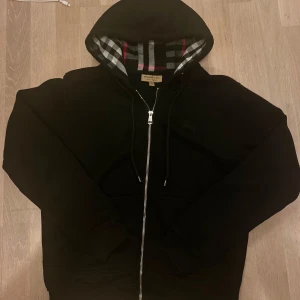  Burberry zip - Svart hoodie från Burberry med klassiskt rutigt foder i huvan och broderad logga på bröstet. Tröjan har hel dragkedja, snörning i huvan och är tillverkad i mjuk bomull. Perfekt för dig som gillar stilrena och exklusiva plagg. Passar S