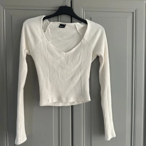 Vit ribbad långärmad topp Gina Tricot - Snygg vit ribbad topp från Gina Tricot med v-ringning och lång ärm. Modellen är croppad och har en figurnära passform som framhäver siluetten. Perfekt att styla med jeans eller kjol för en trendig look.