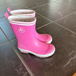 Rosa gummistövlar från McKinley - Snygga rosa gummistövlar från McKinley med vita detaljer och logga på sidan. Perfekta för regniga dagar och äventyr utomhus. Hög modell med praktiska dragflikar upptill och slitstark sula. Färg: fuchsia/white.