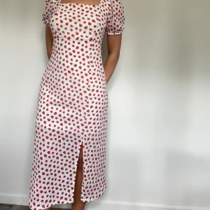 Vit jordgubbsmönstrad klänning Only - Supersöt vit långklänning från Only med röda jordgubbar över hela tyget. Klänningen har korta puffärmar, fyrkantig ringning, slits framtill och öppen rygg med knyt. Perfekt för sommarens alla tillfällen!