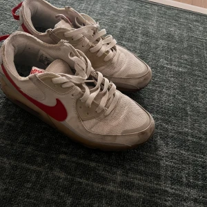 Nike x Parascape vita sneakers med röd swoosh - Nike x Parascape sneakers i vitt mesh och skinn med röd swoosh och detaljer. Låg modell med snörning, beige inslag och genomskinlig sula. Tungan har Parascape-tryck och hälen har röd ögla. Perfekta för dig som gillar unika collabs.