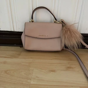Ljusrosa Michael Kors handväska - Söt ljusrosa handväska från Michael Kors med guldfärgade detaljer och en fluffig beige nyckelring. Väskan har både handtag och axelrem. Tillverkad i skinn med stilren design och logga framtill. Den har blovit färgad av mina svarta jens på baksidan, detta har blivit förut och då har jag lyckats få bort de mästa men nu har de blivit igen. Om de önskas kan jah försöka få bort det innan jag skickar om man inte vill gö de själv. Fluffbollen har även blivit lite solblekt på ena sidan. 