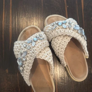 Beige sandaler med pärl- och stendekor - Snygga sandaler i beige med grovt virkat band över foten och glittrande stenar och pärlor som dekoration. Platt sula med liten platå och öppen tå. Perfekta för sommarens alla tillfällen när du vill sticka ut lite extra.