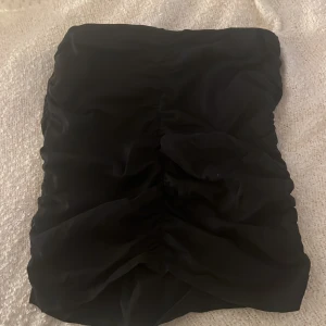 Svart tight kortkjol med rynk - Snygg svart kortkjol med tight passform och rynkade detaljer framtill. Kjolen har en enkel och stilren look som passar perfekt till en trendig outfit. Mjuk och stretchig känsla för extra komfort.