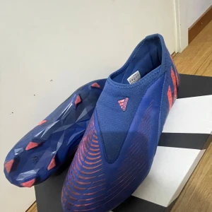 Adidas Predator edge 1  blå fotbollsskor strl 38,5 - Säljer ett par Adidas Predator fotbollsskor i blått med orangea detaljer och mönster. Skorna har en strumpliknande passform utan snörning och är tillverkade i syntetmaterial. Perfekta för dig som vill ha grym kontroll på planen och snygg stil. De är i storlek 38,5 och jag säljer de för jag slutade spela fotboll ungefär när ja köpte de. De är lite svarta pga att jag spelade på konstgräs med de.
