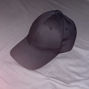 Svart klassisk keps med justerbar rem - Snygg svart keps med böjd skärm och sex paneler. Kepsen har en metallspänne baktill för enkel justering av storlek. Stilren och enkel design utan logga eller mönster, perfekt för dig som gillar minimalistiskt. Materialet känns som bomull eller polyester.