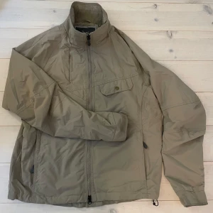 Beige vindjacka från T&P, storlek L - Säljer en  grön vindjacka från T&P i storlek L. Jackan har dragkedja framtill, en bröstficka med knapp och två sidofickor. Materialet är lätt och passar perfekt för utomhusaktiviteter. Snygg och enkel design som funkar till det mesta.
