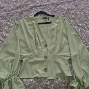 Ljusgrön blus med v-ringning från ASOS - Superfin ljusgrön blus från ASOS DESIGN med v-ringning, tre knappar framtill och ballongärmar med resår vid handlederna. Blusen har en croppad passform och är gjord i ett mjukt, lätt material som ger en flowy känsla.