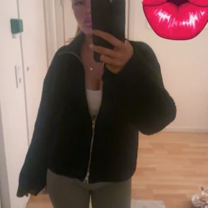 Svart teddyjacka med dragkedja - Mysig svart teddyjacka med hel dragkedja framtill och krage. Jackan har en loose passform och långa ärmar, perfekt för att hålla sig varm och samtidigt se chill ut. Passar dig som gillar en enkel och stilren look.