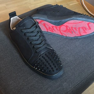 Christian Louboutin svarta sneakers med nitar - Säljer ett par svarta sneakers från Christian Louboutin med ikonisk röd sula och coola svarta nitar på tån. Skorna har snörning och är tillverkade i skinn och mocka. Perfekta för dig som vill sticka ut med en lyxig och edgy stil.
