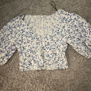 Blommig croppad blus från H&M - Superfin croppad blus från H&M i linneblandning. Vit botten med blått blommönster, v-ringad och med puffiga trekvartsärmar. Perfekt för dig som gillar romantisk och somrig stil. Säljes i storlek S.