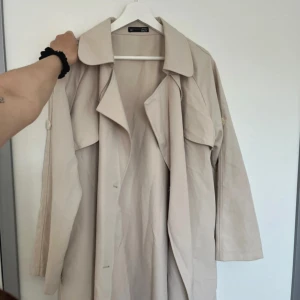 Beige trenchcoat från shein - Stilren beige trenchcoat från shein med klassisk krage och långa ärmar. Jackan har en avslappnad passform och snygga detaljer som breda slag och knappar. Perfekt för lager-på-lager och ger en clean look till din outfit.