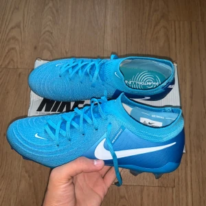 Nike Phantom Luna blå fotbollsskor - Säljer ett par Nike Phantom Luna fotbollsskor i en fräsch blå färg med vita detaljer och snörning. Skorna har en stickad ovandel för extra komfort och passform, samt en platt sula med dobbar för bästa grepp på planen. Perfekta för dig som vill sticka ut på matchen!