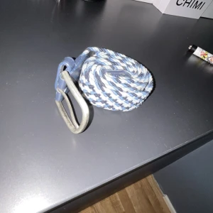 flätat bälte  - Säljer ett snyggt flätat bälte med randigt mönster i blått, vitt och grått. Bältet har ett coolt metallspänne och detaljer i blått läder. Perfekt accessoar för att lyfta din outfit med lite färg och stil.