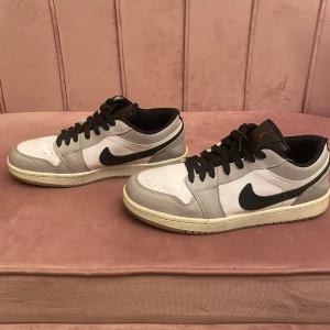 Nike Air Jordan 1 Low grå/vit - Snygga Nike Air Jordan 1 Low sneakers i grått och vitt med svarta detaljer och svart swoosh. Skorna har svart snörning, perforerad tå och klassisk Air Jordan-logga i rött på hälen. Ovandelen är i skinn och de har en platt sula för skön streetstil.