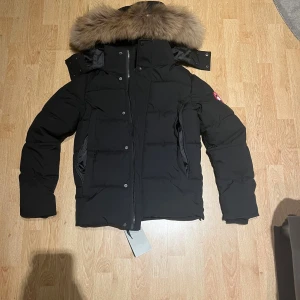 Canada Goose jacka XS - Säljer en svart dunjacka från Canada Goose i storlek XS. Jackan har en fluffig pälskant på huvan, klassisk logga på ärmen och dragkedja med knappar framtill. Perfekt för kalla vinterdagar och riktigt snygg streetstil.
