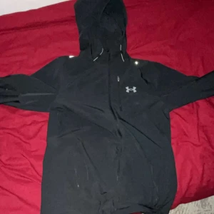Svart vindjacka från Under Armour - Svart vindjacka från Under Armour med huva och dragkedja framtill. Jackan har en stilren look med logga på bröstet och reflekterande detaljer vid axlarna. Perfekt för dig som gillar sportig stil och vill ha något lätt att slänga på sig.