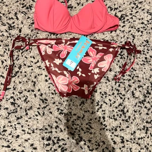 Rosa och vinröd bikini från Scampi - Snygg bikini från Scampi med rosa vadderad bygel-bh och vinröda trosor med stora blommor i rosa och vitt. Trosorna har knyt i sidorna för perfekt passform. Perfekt för stranden eller poolen! Aldrig andvända och har fortfarande lappen kvar då jag köpte i fel storlek💕