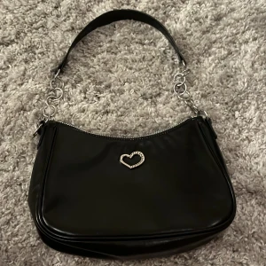 Svart axelväska med hjärtdetalj - Snygg svart axelväska i syntetmaterial med silverfärgad dragkedja och kedjedetaljer på axelbandet. Framsidan har en liten hjärtformad metallapplikation som ger väskan en söt touch. Perfekt storlek för det viktigaste och enkel att matcha med olika outfits. Aldrig använd 
