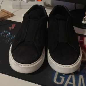 Svarta slip-on sneakers med vit sula - Säljer ett par svarta slip-on sneakers med tjock vit sula. Skorna har en minimalistisk design utan synlig snörning och är tillverkade i mocka och skinn. Perfekta för dig som gillar stilrena och enkla skor med modern vibe.
