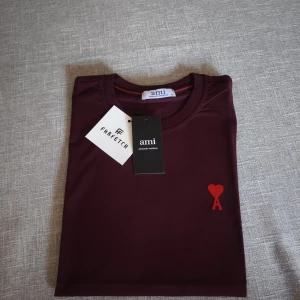 Vinröd Ami Paris T-shirt  - Vinröd t-shirt från Ami Paris med klassisk passform och rund hals. På bröstet finns Ami:s ikoniska röda hjärta med bokstaven A broderat. T-shirten är i mjuk bomull och har korta ärmar.