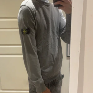 Grå Stone Island sweatshirt - Säljer en grå sweatshirt från Stone Island Junior med klassisk patch på vänster ärm. Tröjan har rund hals, ribbade muddar och en clean look som passar till jeans eller joggers. Perfekt för dig som gillar streetwear och vill ha något stilrent.Passar mig bra och jag är 180 och väger 60 kg med dragkedjor på sidorna av magen