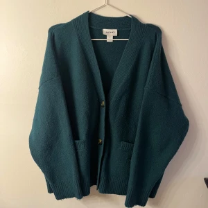 Kofta - Säljer en oversized mörkgrön kofta från Monki med djup v-ringning och stora knappar framtill. Koftan har två fickor och breda ribbade muddar vid ärmslut och nederkant. Perfekt att slänga på sig när det är kyligt ute.