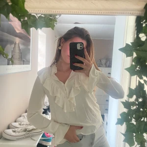 Vit blus Zara med volangdetaljer - Säljer en superfin vit blus med volang längs v-ringningen och framtill. Blusen har långa ärmar och en luftig passform som ger en romantisk vibe. Perfekt att styla med jeans eller kjol för en trendig look.