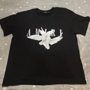 Svart AMIRI t-shirt med duvor - Svart t-shirt från AMIRI med stort tryck av två vita duvor och rosa bokstäver på bröstet. Klassisk passform och rund halsringning. Perfekt för dig som vill sticka ut med en snygg och modern streetstyle.