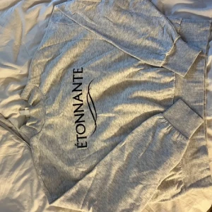 Grå sweatshirt med Étonnante-tryck - Snygg grå sweatshirt med svart Étonnante-tryck på bröstet. Tröjan har rund hals, långa ärmar och breda muddar vid ärmslut och nederkant. Perfekt för en chill och stilren look.